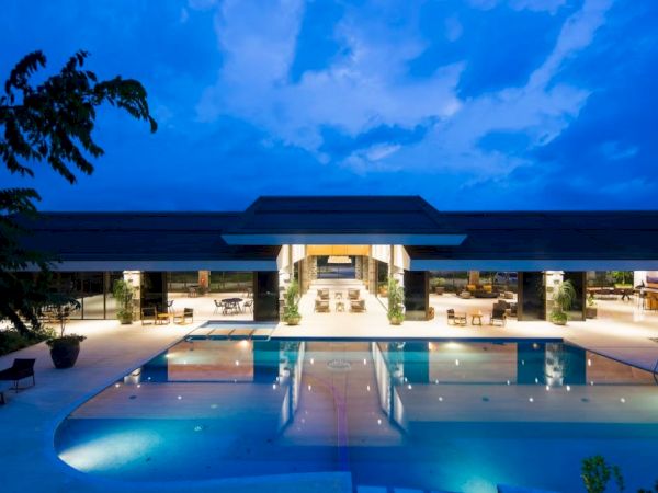 A resort at night with a large swimming pool, loungers, and an open-air pavilion, illuminated under a deep blue sky.