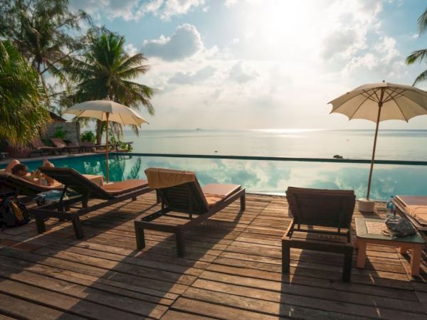 An inviting poolside with lounge chairs, umbrellas, a wooden deck, and a stunning view of the sea under a partly cloudy sky and palm trees.