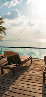 A serene poolside view with lounge chairs, umbrellas, palm trees, and a tranquil waterbody reflecting the cloudy sky at sunrise or sunset.