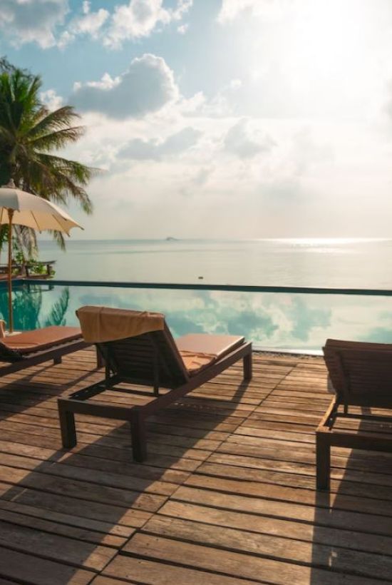 The image showcases a serene poolside with lounge chairs, umbrellas, and a view of the ocean against a partly cloudy sky, with palm trees in the background.