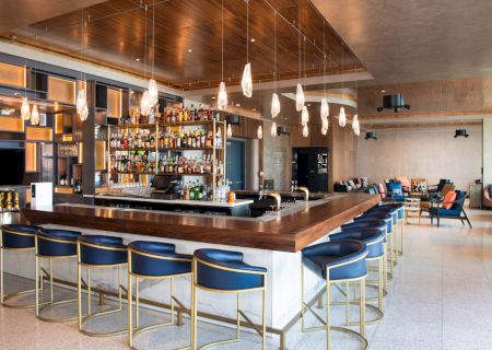 A modern bar with sleek blue stools, hanging pendant lights, a well-stocked liquor shelf, and a cozy seating area in the background.