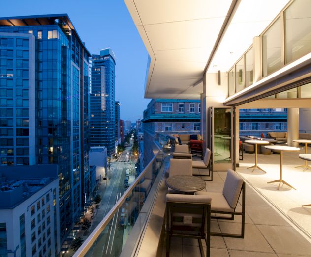 A modern cityscape view from a high-rise balcony at dusk, featuring outdoor seating and tables, with buildings and a street visible below.