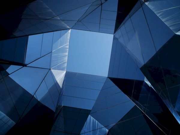 Looking up through a geometric arrangement of reflective, angular glass structures, revealing a portion of clear blue sky at the center.