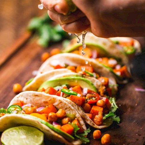 A hand is squeezing lime juice over a row of freshly prepared tacos filled with vegetables, chickpeas, avocado slices, and garnished with cilantro.