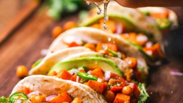 A hand is squeezing lime juice over a row of freshly prepared tacos filled with vegetables, chickpeas, avocado slices, and garnished with cilantro.