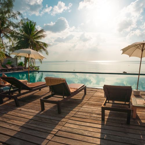 Lounge chairs and umbrellas on a wooden deck overlook a serene infinity pool with a tropical ocean view under a bright sky.