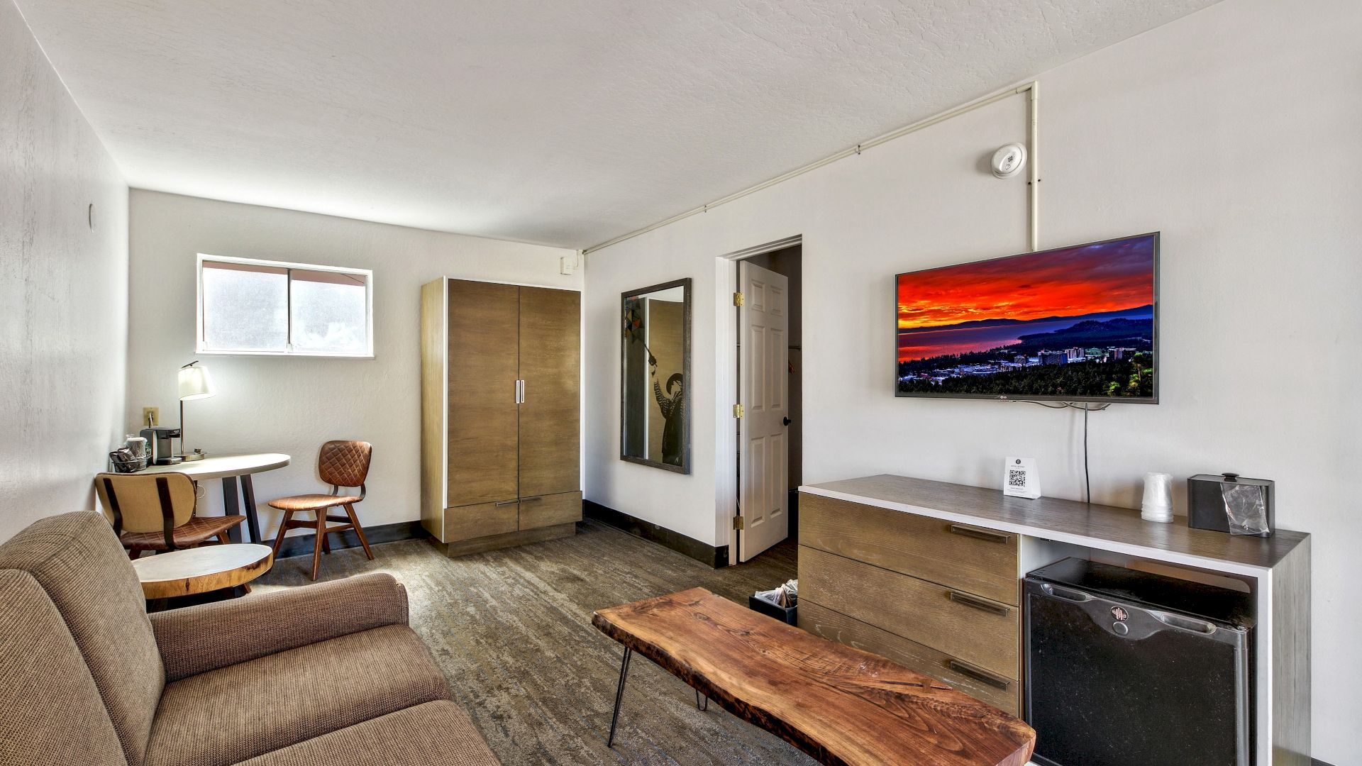 A modern room featuring a brown couch, a wooden coffee table, a TV displaying a sunset, a wardrobe, a desk with a chair, and a mini fridge.