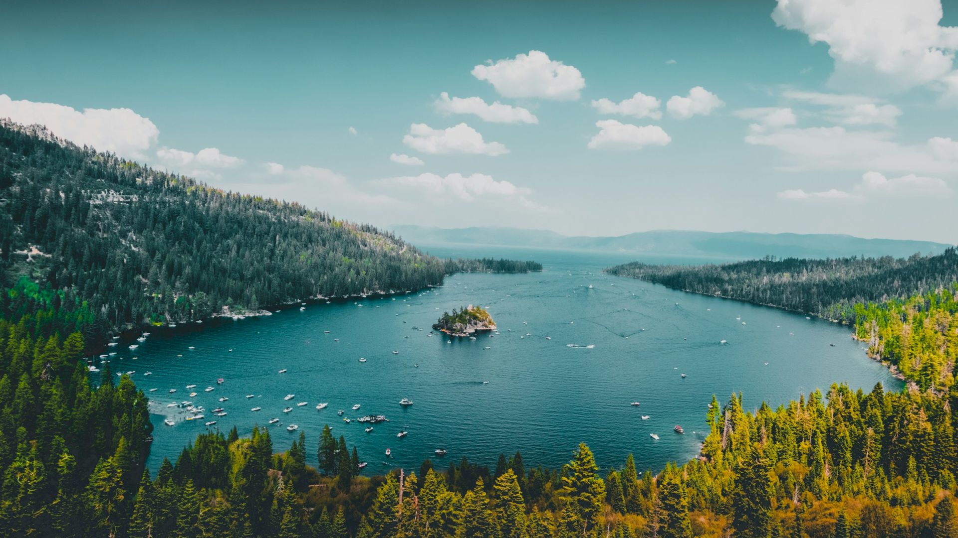 A picturesque lake surrounded by lush green forests, with an island in the center and mountains in the distance under a partly cloudy sky.
