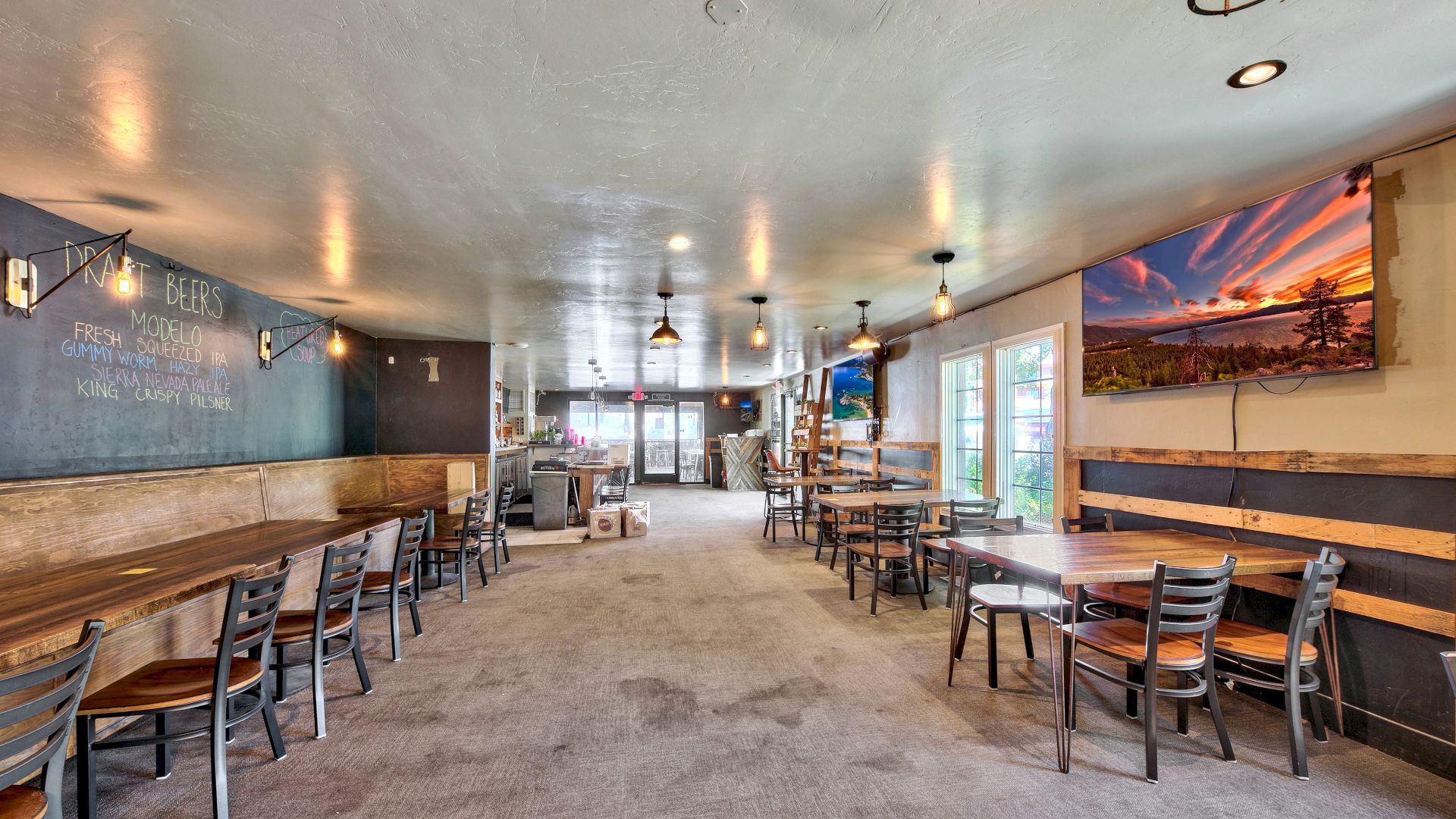 The image shows the interior of a spacious and well-lit cafe or restaurant with wooden tables and chairs, wall art, and a large window.