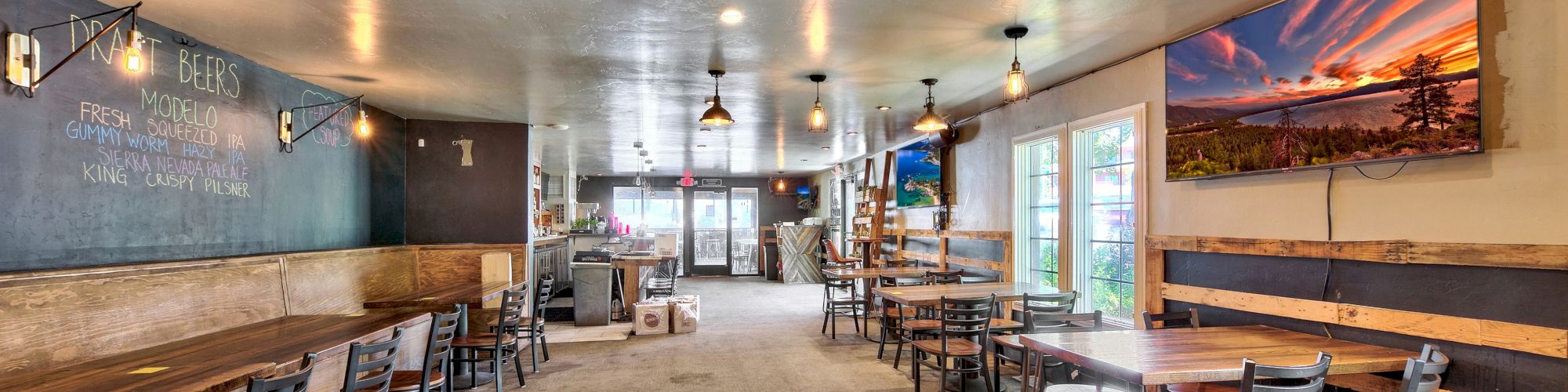 The image shows the interior of a spacious and well-lit cafe or restaurant with wooden tables and chairs, wall art, and a large window.