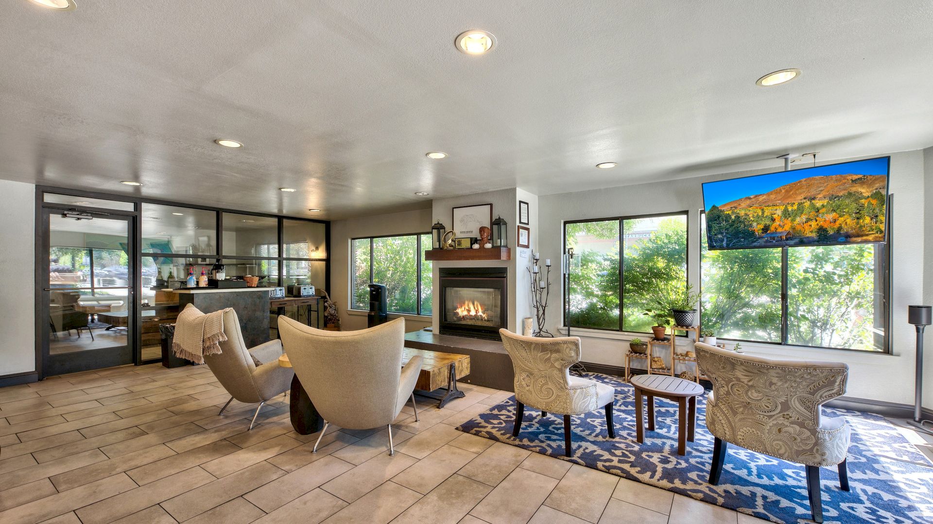 A modern living room features beige chairs, a fireplace, and large windows with scenic views. There's a blue patterned rug and a dining area.
