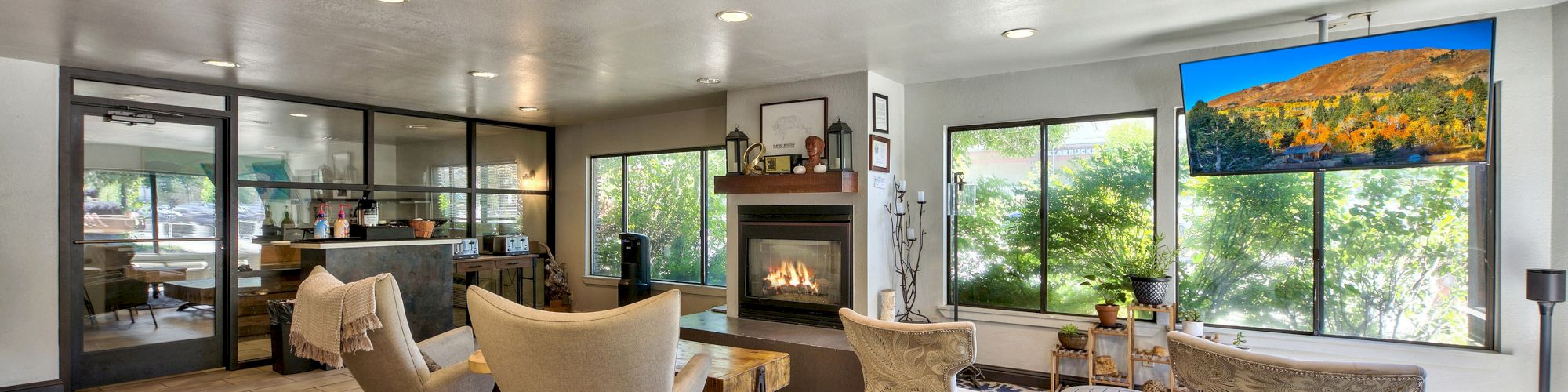 A modern living room features beige chairs, a fireplace, and large windows with scenic views. There's a blue patterned rug and a dining area.