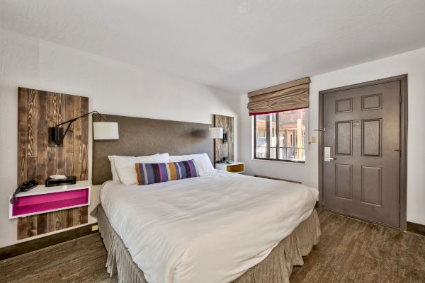 The image shows a neatly made bed in a hotel room with bedside tables, a window with blinds, and a wooden door.