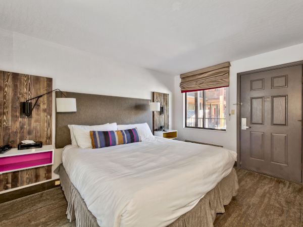 The image shows a neatly made bed in a hotel room with bedside tables, a window with blinds, and a wooden door.