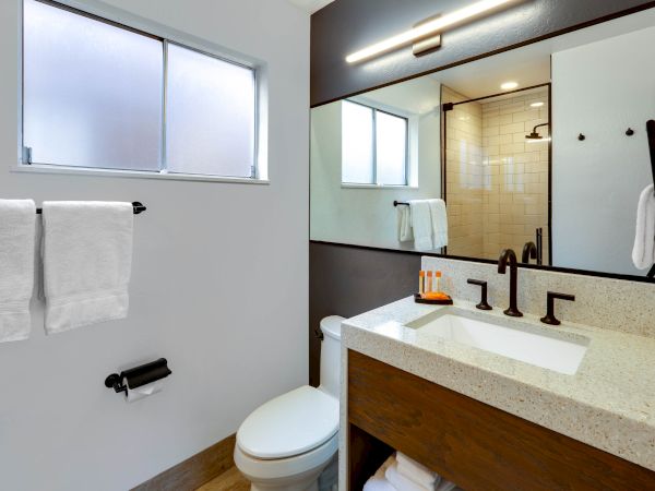 A modern bathroom with a large mirror, white countertop, black faucet, toilet, towels, and a glass-enclosed shower with subway tiles.