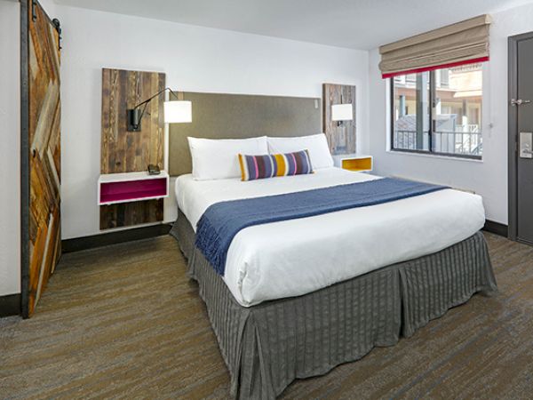 A well-lit hotel room with a neatly made bed, colorful pillows and blanket, wall-mounted lamps, rustic nightstands, window, and a door.