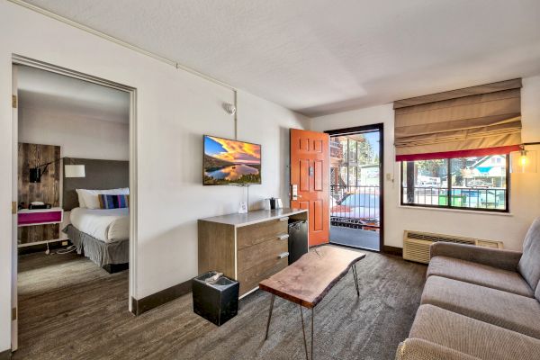 A furnished living room with a couch, coffee table, TV, and an open door leading to a balcony; bedroom visible through an adjacent doorway.