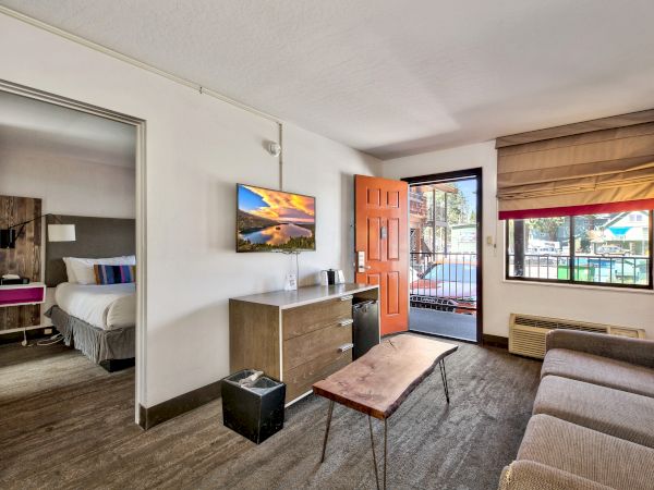 A furnished living room with a couch, coffee table, TV, and an open door leading to a balcony; bedroom visible through an adjacent doorway.