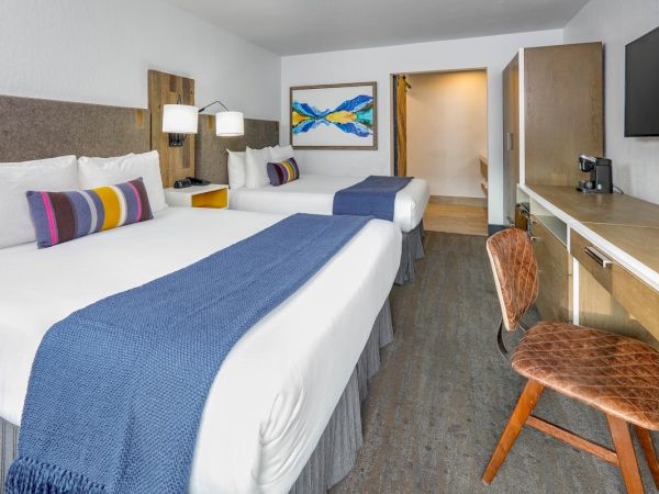 A hotel room with two beds, blue blankets, colorful pillows, a desk, chair, TV, and wall art in a modern minimalist style ending the sentence.