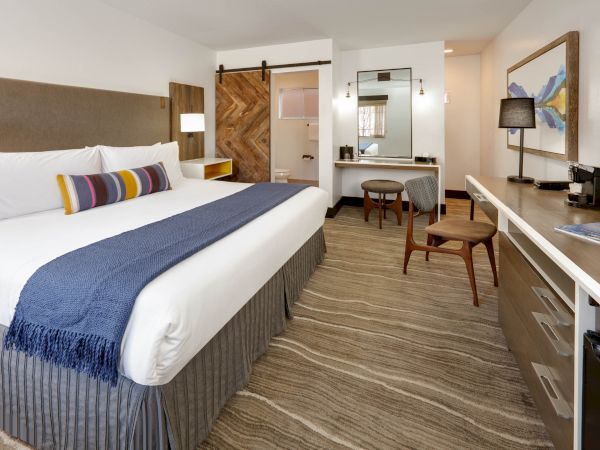 A modern hotel room with a king-sized bed, blue throw, wood accents, desk, mirror, coffee maker, and a sliding barn door leading to the bathroom.