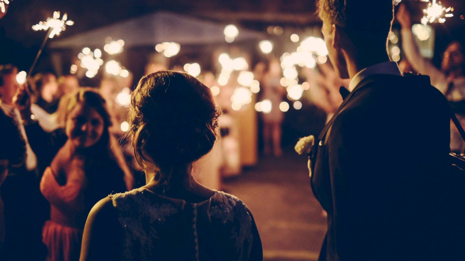 A couple is standing with their back to the camera, surrounded by people holding sparklers, creating a festive and celebratory atmosphere.