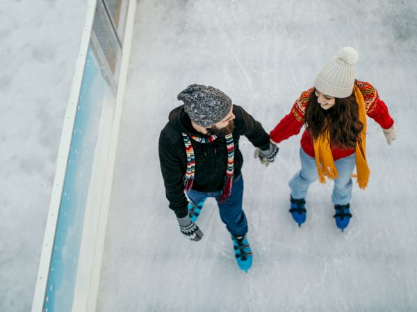 Two people are ice skating holding hands, both wearing winter clothing including hats and scarves, with a barrier on the left.