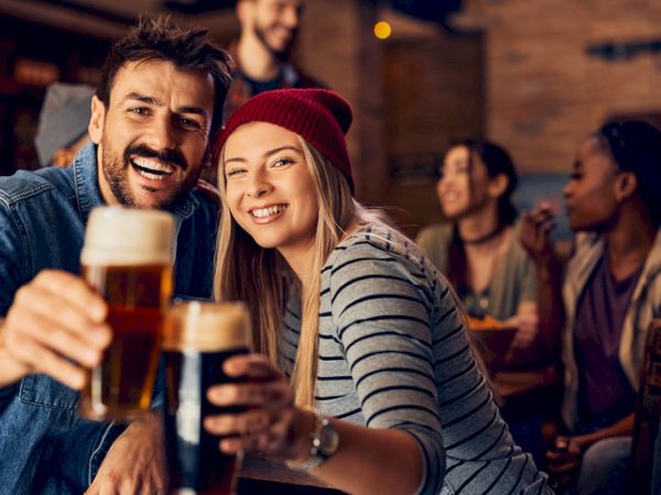 Two people hold up glasses of beer, smiling at the camera in a lively social setting with friends in the background, enjoying their time together.
