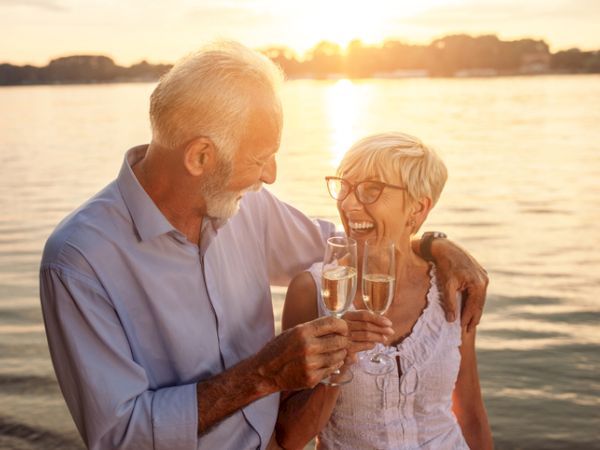 An older couple is enjoying a sunset by the water, smiling and toasting with champagne glasses, exuding happiness and love.