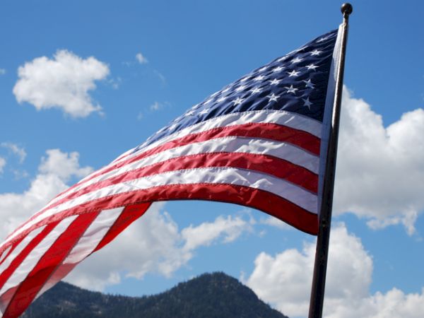 An American flag is waving in the wind with a clear blue sky and a mountainous landscape in the background.