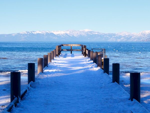 A snow-covered wooden pier stretches out into a calm, blue lake with mountains in the background under a clear sky, creating a serene scene.
