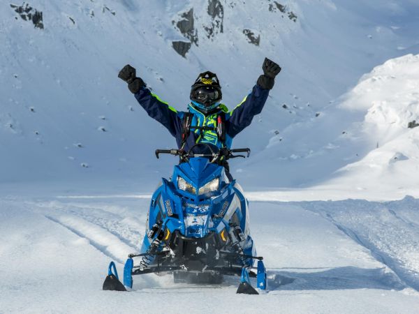 A person riding a blue snowmobile is raising their arms triumphantly on a snowy landscape with mountains in the background.