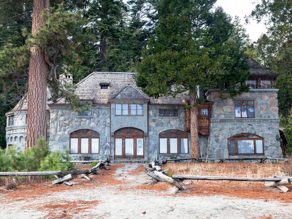 A large stone house surrounded by trees and a dirt path leading to it. The building has multiple windows and a rustic appearance.