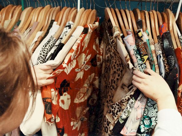 A person is browsing through a rack of various colorful clothes on wooden hangers, with patterns such as florals and animal prints.