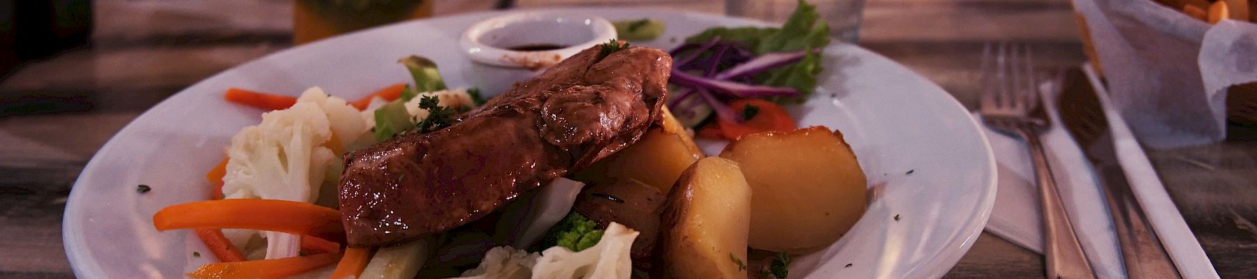 A plate of grilled meat with mixed vegetables and potatoes, a drink with orange slices, a glass of water, fries, and utensils on a wooden table.