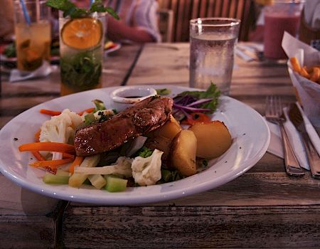 A plate of grilled meat with mixed vegetables and potatoes, a drink with orange slices, a glass of water, fries, and utensils on a wooden table.