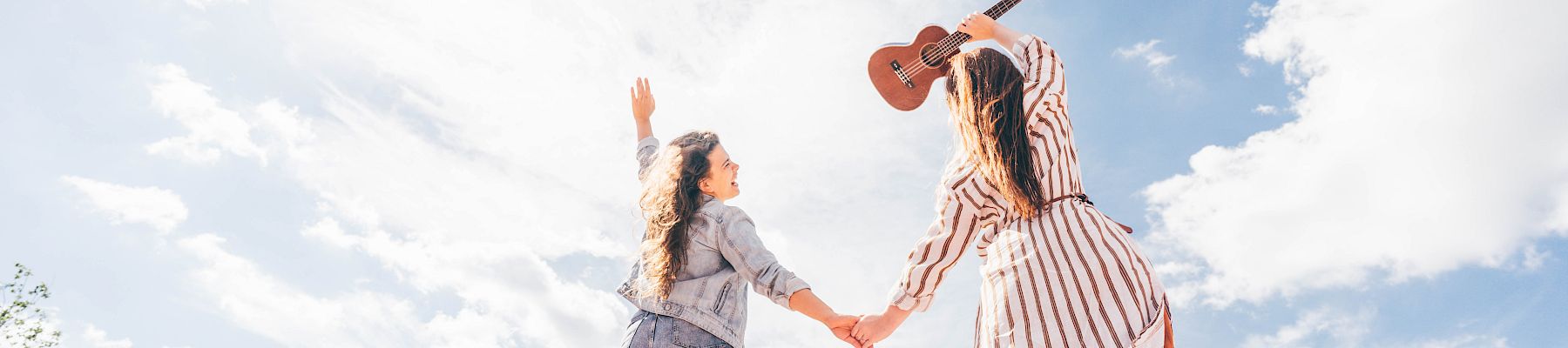 Two people holding hands outdoors, one with a guitar, both reaching toward the sky, under a bright sunny day with scattered clouds, trees in the background.