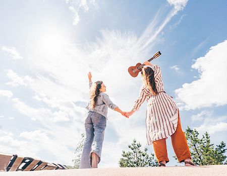 Two people holding hands outdoors, one with a guitar, both reaching toward the sky, under a bright sunny day with scattered clouds, trees in the background.