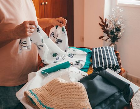 A person is packing a suitcase with various clothing items, including a patterned shirt, a knitted item, and some folded clothes, standing in a room.