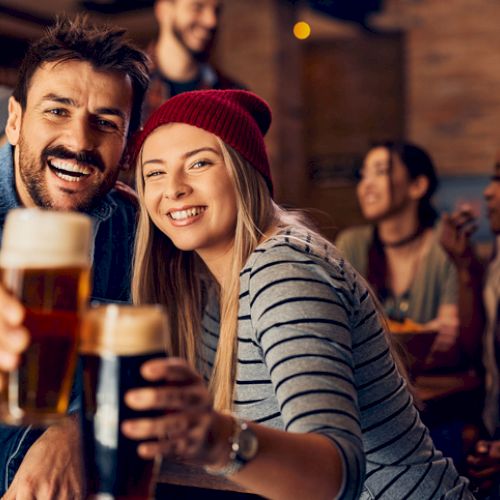 A group of friends is enjoying their time at a bar, with two people in the foreground smiling and holding up glasses of beer.