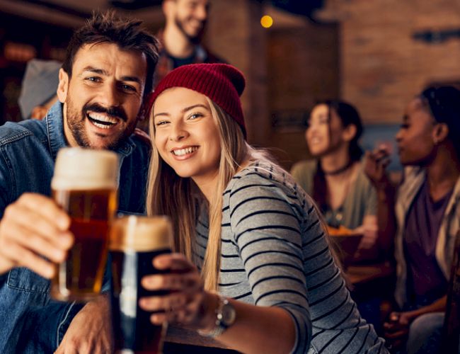 A group of friends is enjoying their time at a bar, with two people in the foreground smiling and holding up glasses of beer.