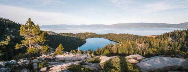 A serene mountainous landscape featuring a clear blue lake surrounded by lush green forests and rocky terrain under a blue sky with wispy clouds.