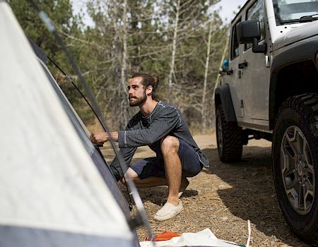 A man is setting up a tent in a wooded area next to a parked SUV, preparing for a camping trip.