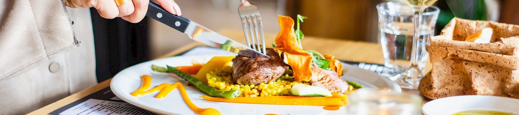 A person is eating a meal with a knife and fork, with a dish of colorful vegetables and meat, alongside wine, bread, and a candle on the table.