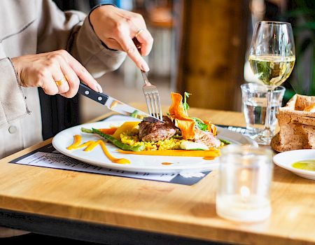 A person is eating a meal with a knife and fork, with a dish of colorful vegetables and meat, alongside wine, bread, and a candle on the table.