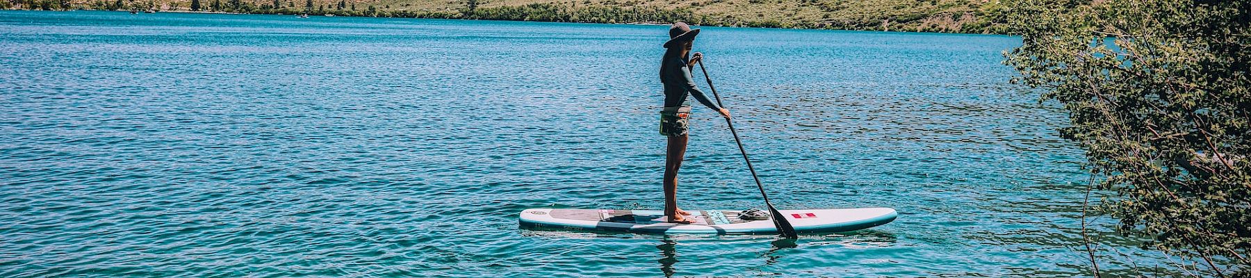 A person is paddleboarding on a clear, blue lake surrounded by hills with greenery under a blue sky.