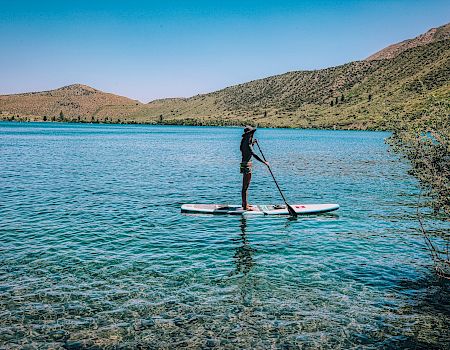 A person is paddleboarding on a clear, blue lake surrounded by hills with greenery under a blue sky.