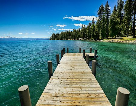 A wooden pier extends into a clear blue lake surrounded by tall pine trees on a sunny day, with mountains visible in the distance.