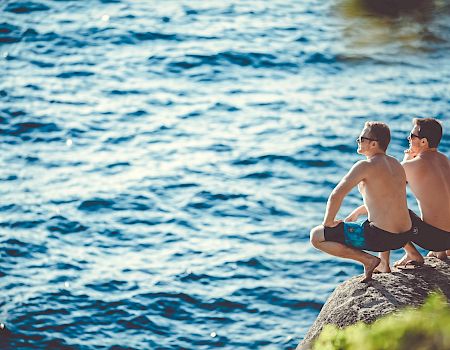 Two shirtless individuals are sitting on a rock by a body of water, looking out at the water, under a sunny sky, with greenery nearby.