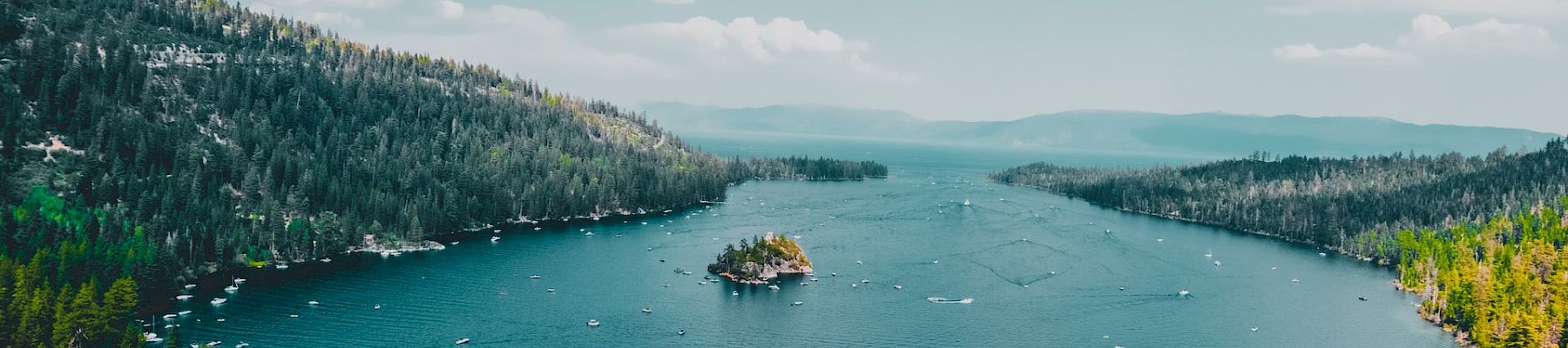 A serene lake surrounded by dense forests and mountains, with boats scattered on the water and a small island in the center under a partly cloudy sky.