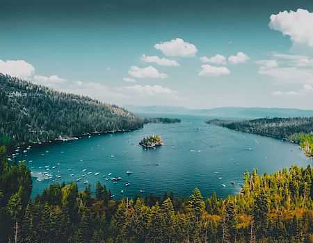 A serene lake surrounded by dense forests and mountains, with boats scattered on the water and a small island in the center under a partly cloudy sky.